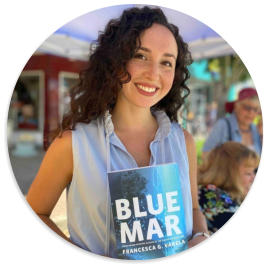 Francesca Varela wearing a blue dress and holding up a copy of her book, Blue Mar