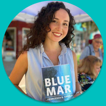 Francesca Varela wearing a blue dress and holding up a copy of her book, Blue Mar